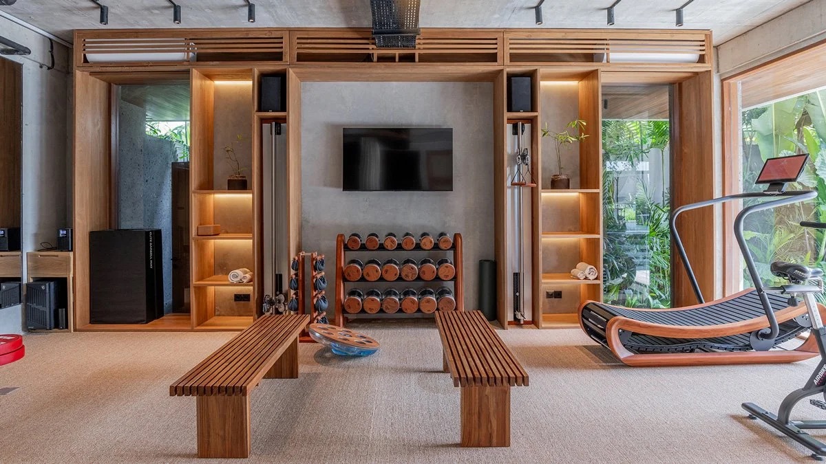 Minimal wood-panelled home gym with dumbbells, bench, treadmill, and a view of tropical plants