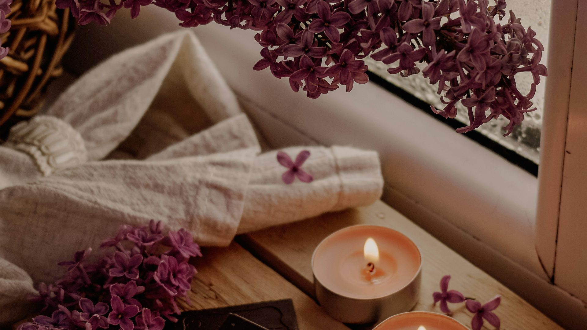 A rolled linen cloth, cascading lilacs, and a lit tea-light candle on a wooden windowsill